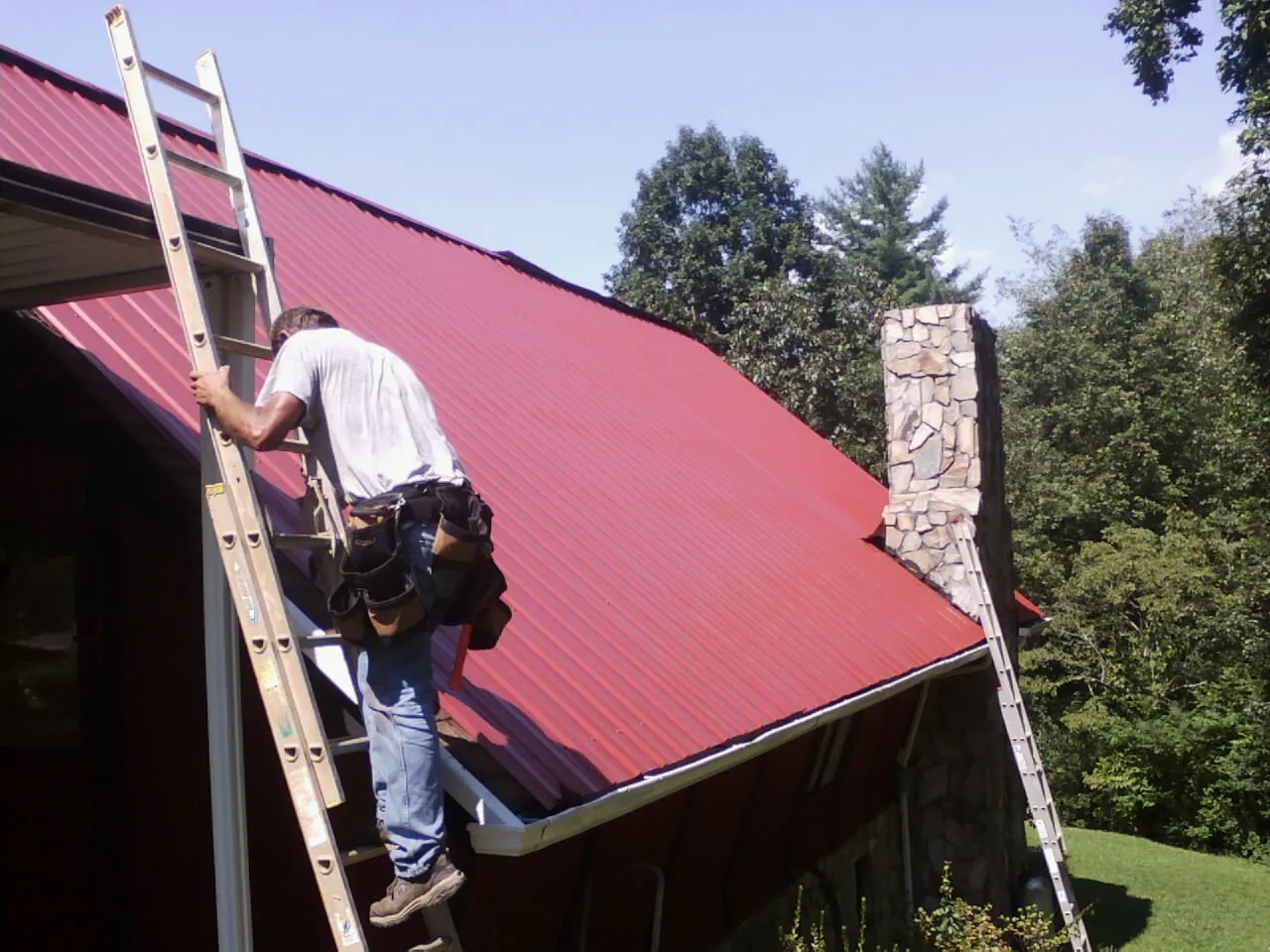 Roofer on a ladder beside a red metal roof and stone chimney — Soffit & Fascia Repair by Roofing Brothers in Statesboro